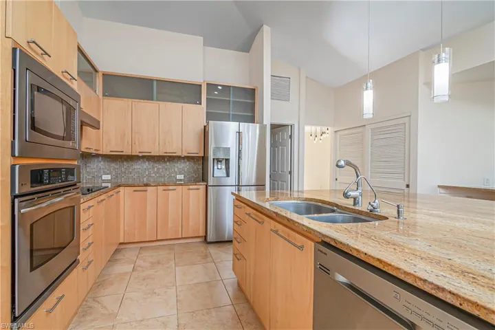 Kitchen with glass insert cabinets, light stone counters, light brown cabinets, appliances with stainless steel finishes, and hanging light fixtures