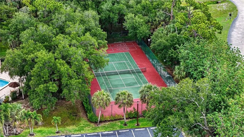 Tennis court with green and red hard surfacing, surrounded by mature trees, privacy fencing, and palm trees