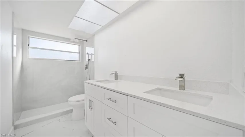 Full bathroom featuring double vanity, a marble finish shower, light marble finish floors, and a skylight