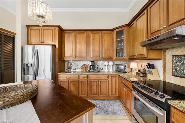 Kitchen featuring glass insert cabinets, appliances with stainless steel finishes, brown cabinets, under cabinet range hood, and pendant lighting