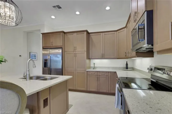 Kitchen featuring stainless steel appliances, light stone counters, wood finish cabinetry, suspended lighting, and ornamental molding