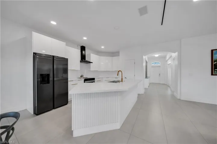 Kitchen with black refrigerator with ice dispenser, white cabinets, arched walkways, a kitchen island with sink, and wall chimney range hood