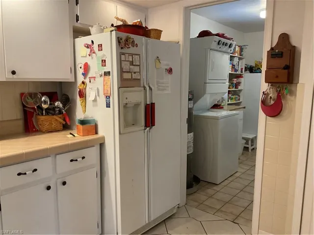 Kitchen featuring tile counters, white cabinetry, white refrigerator with ice dispenser, stacked washer and clothes dryer, and light tile patterned flooring