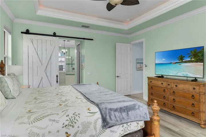 Bedroom with light wood finished floors, ceiling fan, ensuite bath, crown molding, and a tray ceiling