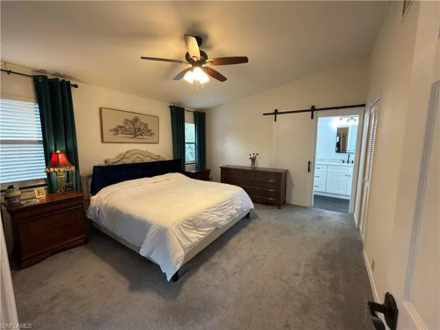 Bedroom with a barn door, vaulted ceiling, carpet floors, a ceiling fan, and ensuite bath