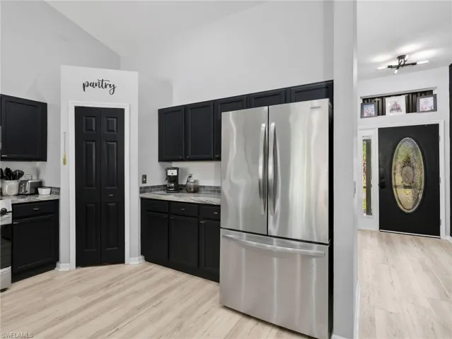 Kitchen with freestanding refrigerator, dark cabinetry, light wood finished floors, vaulted ceiling, and light stone countertops