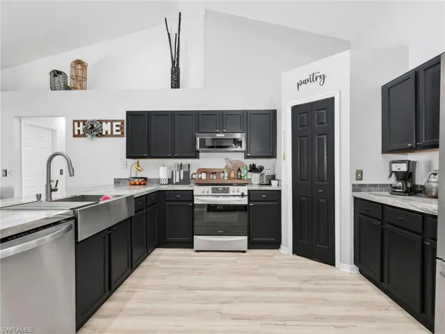 Kitchen with stainless steel appliances, dark cabinets, light stone counters, light wood-type flooring, and vaulted ceiling