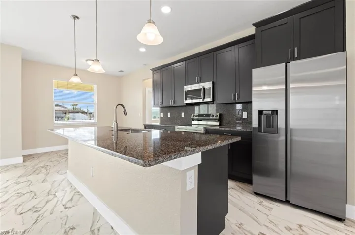 Kitchen with stainless steel appliances, dark stone countertops, pendant lighting, a kitchen island with sink, and light marble finish floors