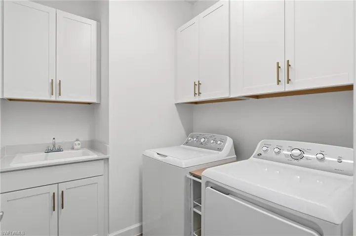 Laundry room featuring white shaker-style cabinetry, a built-in utility sink with a stone-finish countertop, and brass-finish hardware