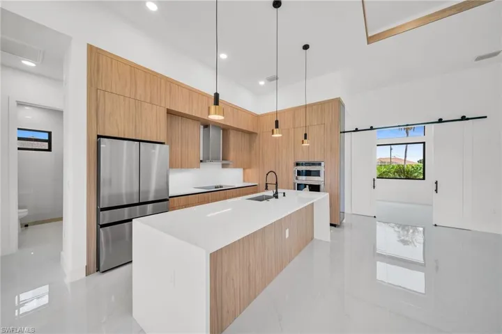 Kitchen with stainless steel appliances, hanging light fixtures, a kitchen island with sink, light tile patterned flooring, and wall chimney exhaust hood
