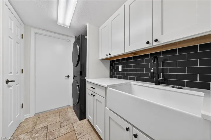 The laundry area features stacked appliances, white cabinetry with black hardware, a farmhouse-style sink with a black faucet, and a black subway tile backsplash