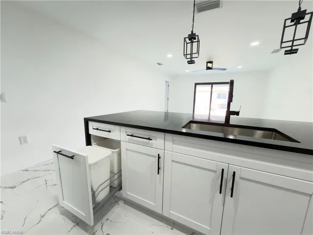Kitchen with dark stone countertops, white cabinetry, decorative light fixtures, and light marble finish floors