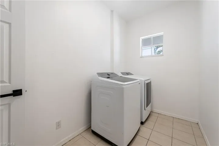 Laundry area featuring light-toned tile flooring, a window providing natural light, and a white paneled door with a matte black handle