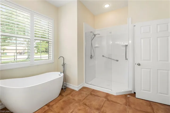 Bathroom featuring a freestanding soaking tub with floor-mounted faucet, a clear glass enclosed shower with grab bars, and large windows with plantation shutters