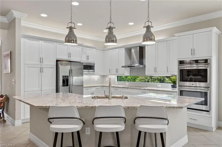 Kitchen with wall chimney range hood, stainless steel appliances, a kitchen island with sink, sink, and light tile floors