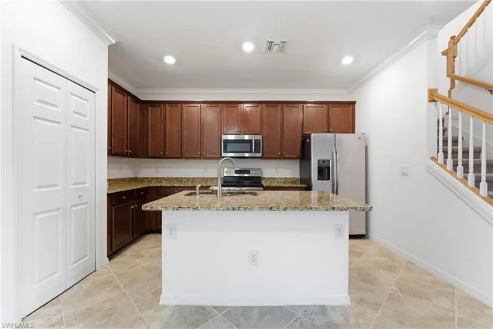 Kitchen featuring stainless steel appliances, light stone counters, a kitchen island with sink, dark wood finish cabinets, and ornamental molding