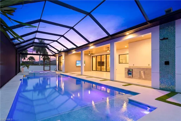 Resort-style screened lanai with a heated pool and spa, sun shelf, outdoor kitchen, covered lounge area, and dramatic waterfall wall - beautifully lit for evening entertaining!