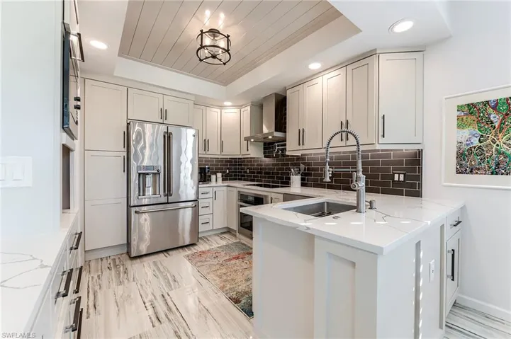 Kitchen featuring a raised ceiling, stainless steel appliances, light stone counters, backsplash, and wall chimney range hood