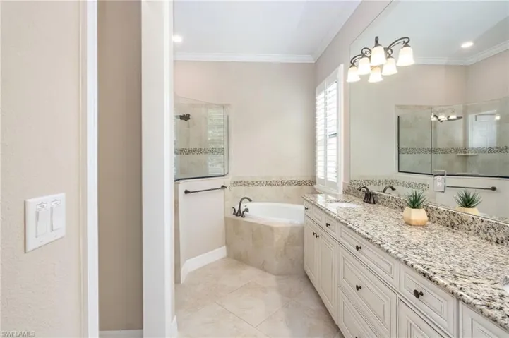 Dual vanity with stone countertop, built-in soaking tub, glass-enclosed shower, plantation shutters, and tiled flooring