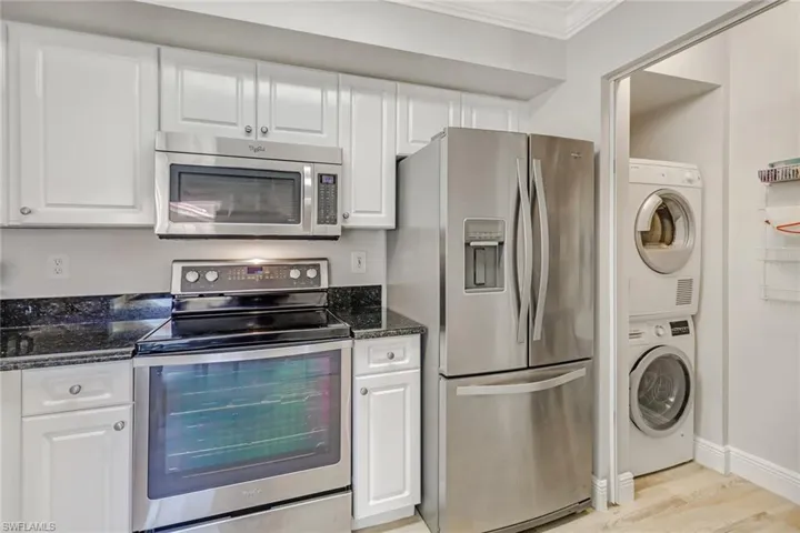 Kitchen featuring stainless steel appliances, white cabinetry, dark stone counters, stacked washer and dryer, and ornamental molding