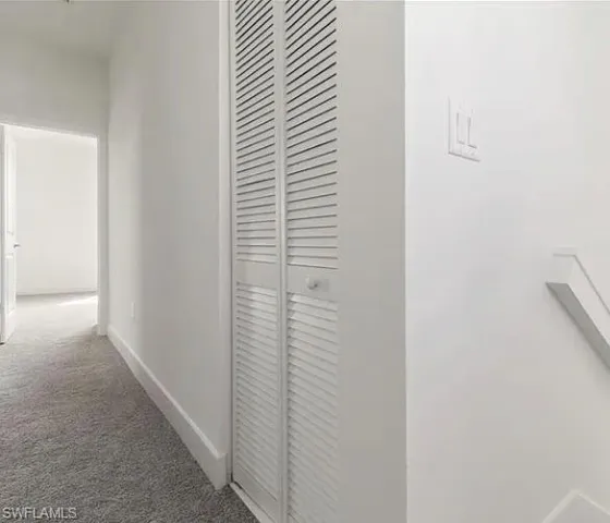 Hallway featuring louvered bi-fold doors, grey carpet, white baseboards, and a doorway leading to an additional space