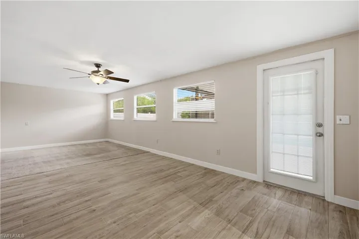 Spacious room featuring wood-finish flooring, light neutral wall tones, multiple windows with horizontal blinds, a ceiling fan with integrated lighting, and a white paneled door with integrated blinds
