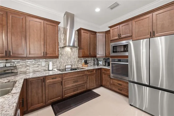 Kitchen with stainless steel appliances, ornamental molding, light stone counters, wall chimney exhaust hood, and backsplash