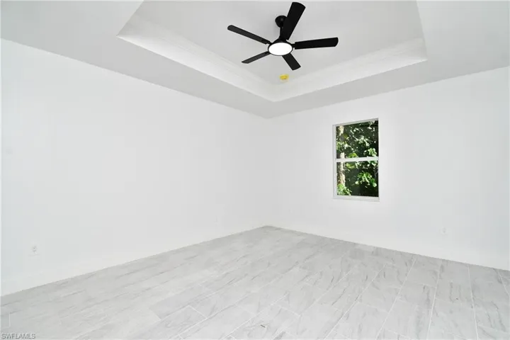 Recessed ceiling with crown molding, ceiling fan with integrated lighting, window with exterior greenery, and light-toned tile flooring