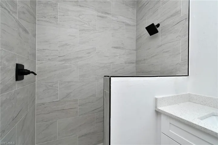 Tiled shower with matte black fixtures, glass shower partition, white vanity with shaker-style door, and speckled countertop