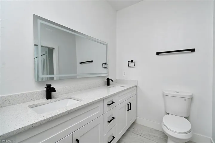 Double vanity with integrated sinks, speckled countertop, white cabinetry with black hardware, back-lit vanity mirror, and modern black fixtures