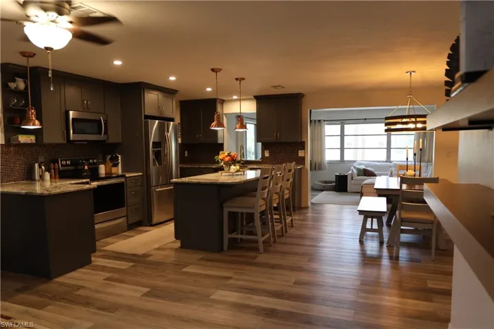 Open-concept kitchen featuring wood-finish flooring, dark cabinetry, stainless steel appliances, a granite-style island with seating, and recessed lighting