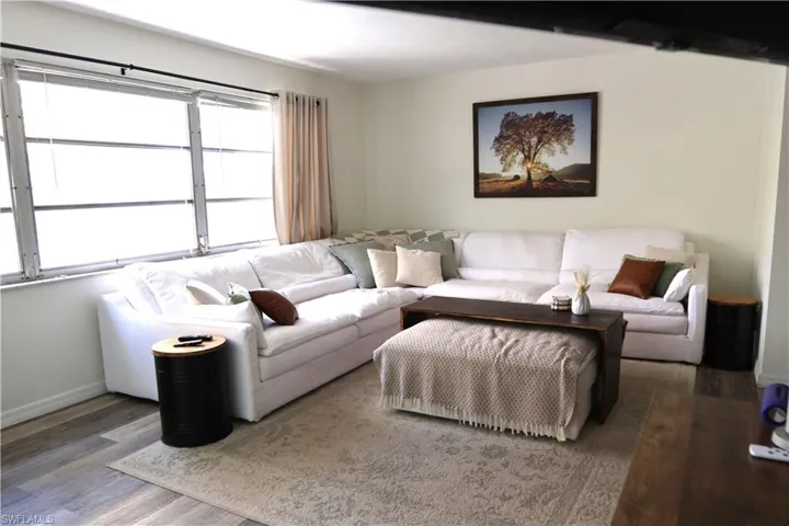 Spacious living area featuring light-toned walls, wood-finish flooring, and a large window with vertical blinds