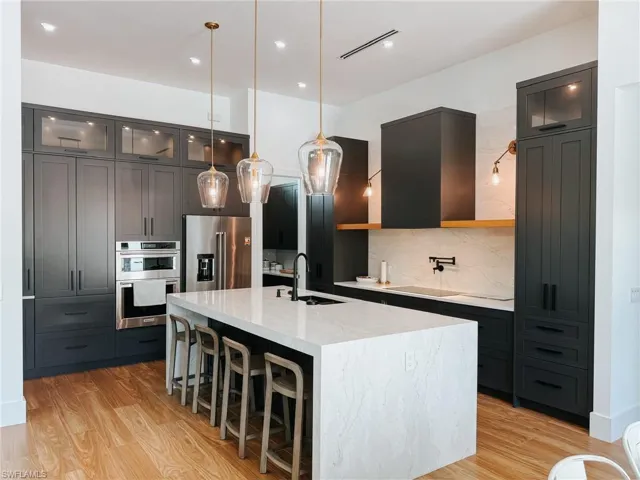 Gourmet kitchen featuring a large waterfall island with integrated sink, dark cabinetry with glass-front uppers, stainless steel appliances, wood-finish flooring, and a white tile backsplash