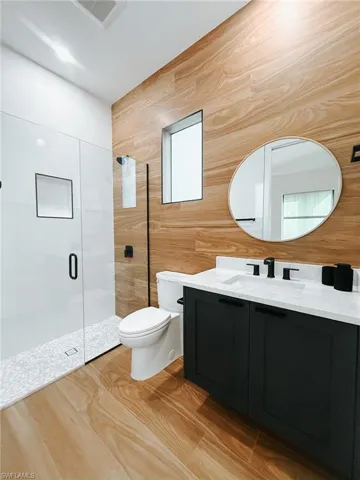 Bathroom featuring wood-finish wall paneling, a dark wood-finish vanity with a white countertop and rectangular sink, matte black fixtures, a frameless oval mirror, and a glass-enclosed shower with pebble-style flooring