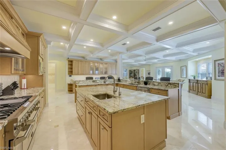 Kitchen with coffered ceiling, beam ceiling, light stone counters, glass insert cabinets, and custom range hood