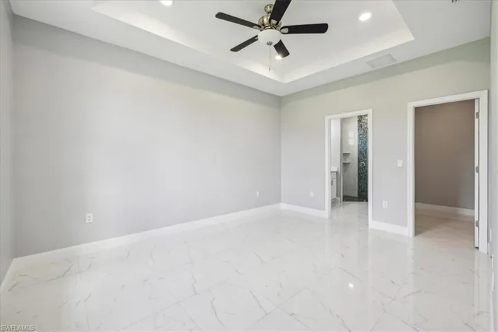 Unfurnished bedroom with light tile patterned flooring, ceiling fan, a tray ceiling, ensuite bath, and a walk in closet
