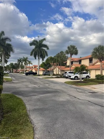 The community features residences with light-colored exteriors and red tile roofs, set along a paved street with green lawns and mature palm trees