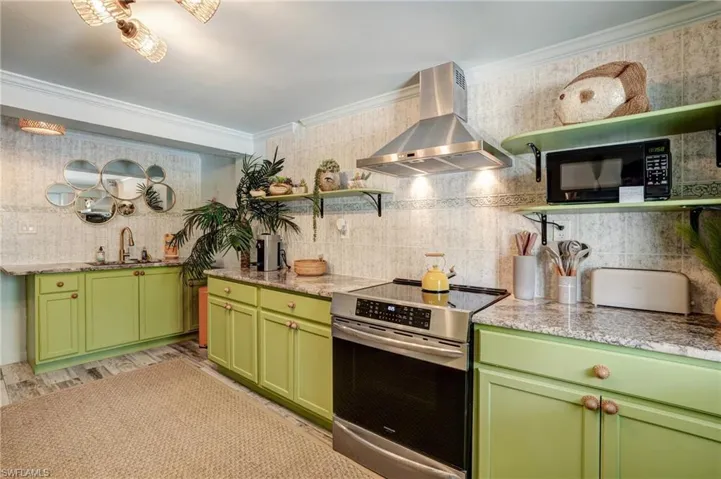 Kitchen with wall chimney range hood, stainless steel electric range, green cabinetry, black microwave, and ornamental molding - Virtually Edited Image