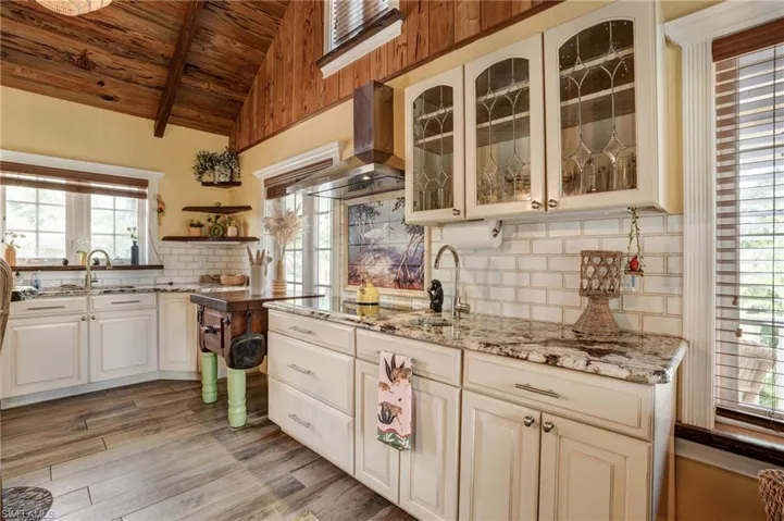 Kitchen featuring wood ceiling, wall chimney range hood, a sink, decorative backsplash, and light stone counters- Virtually Edited Image