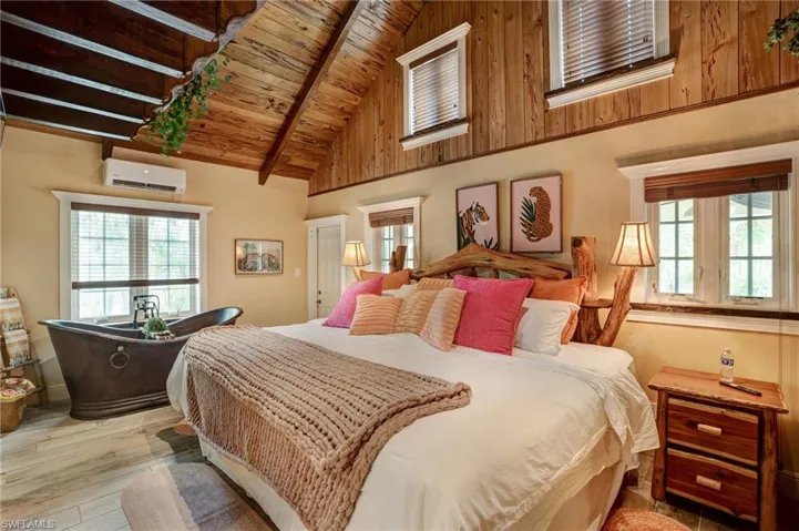 Bedroom with a wall unit AC, wooden ceiling with exposed beams, multiple windows, light wood-type flooring, and high vaulted ceiling- Virtually Edited Image