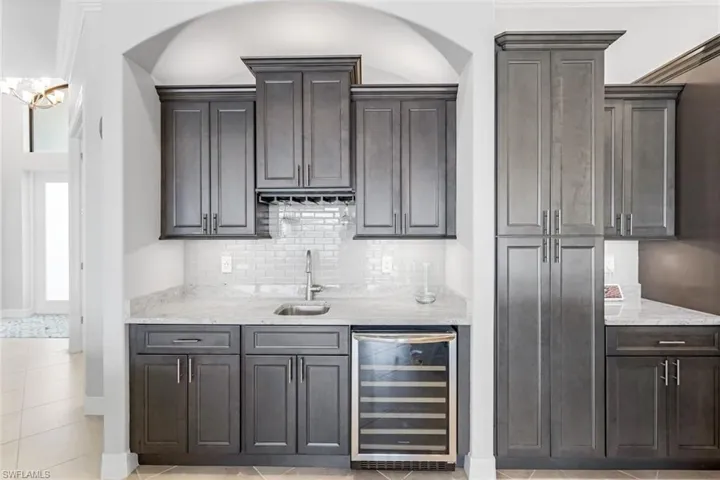 Bar with light stone counters, backsplash, wine cooler, dark brown cabinets, and light tile floors
