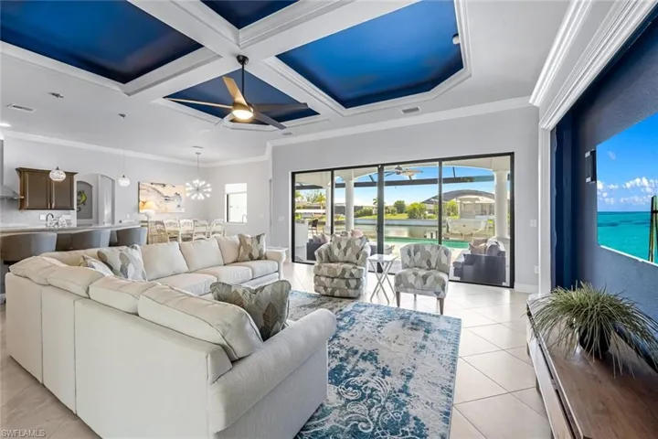 Living room with ornamental molding, light tile flooring, ceiling fan with notable chandelier, and coffered ceiling