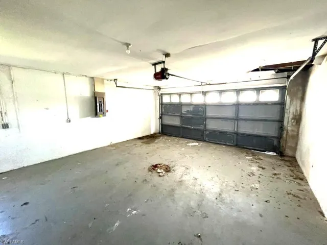 Garage space featuring a paneled roll-up door with upper window sections, an exposed garage door opener, and a wall-mounted electrical panel