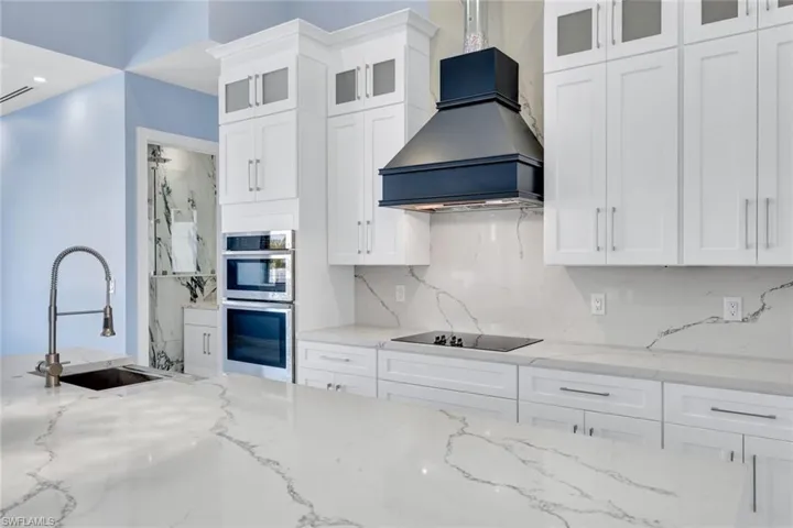 Kitchen with white cabinetry, glass insert cabinets, light stone counters, stainless steel double oven, and backsplash