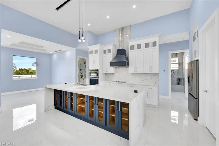 Kitchen featuring glass fronted cabinets, white cabinetry, a large island with sink, light stone countertops, and pendant lighting