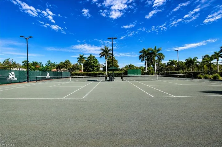Two hard-surface tennis courts featuring perimeter fencing, multiple light poles, a central shaded seating area, and mature palm trees