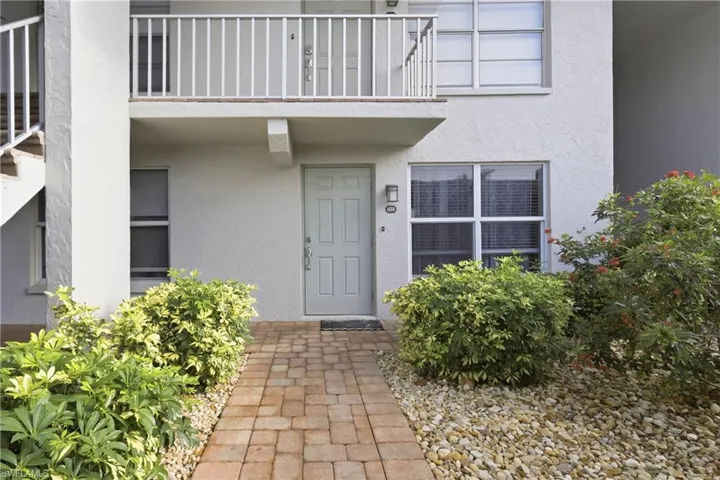 Entryway featuring a paver walkway, mature landscaping, and an upper-level balcony with white railings