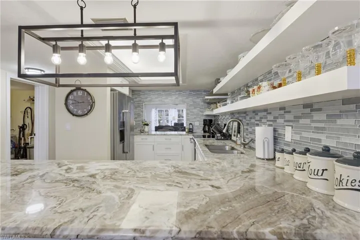 Spacious kitchen featuring a large island with a natural stone countertop, a modern rectangular pendant light fixture, a mosaic tile backsplash, white cabinetry, and stainless steel appliances