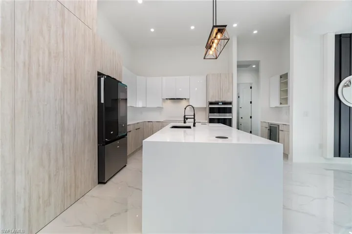 Kitchen featuring stainless steel double oven, a kitchen island with sink, black fridge, hanging light fixtures, and white cabinets