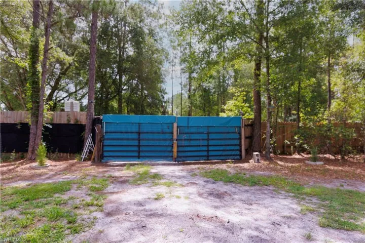 Double-leaf metal gate with a teal covering, set within a natural tree line and partially visible wood fencing